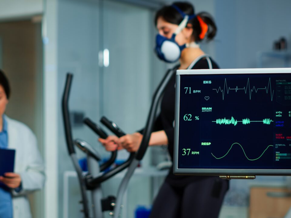 Monitor showing EKG scan of athlete running on cross trainer and medical specialists supervise exercise in background, controlling physical activity, measuring heart rate in sports science laboratory
